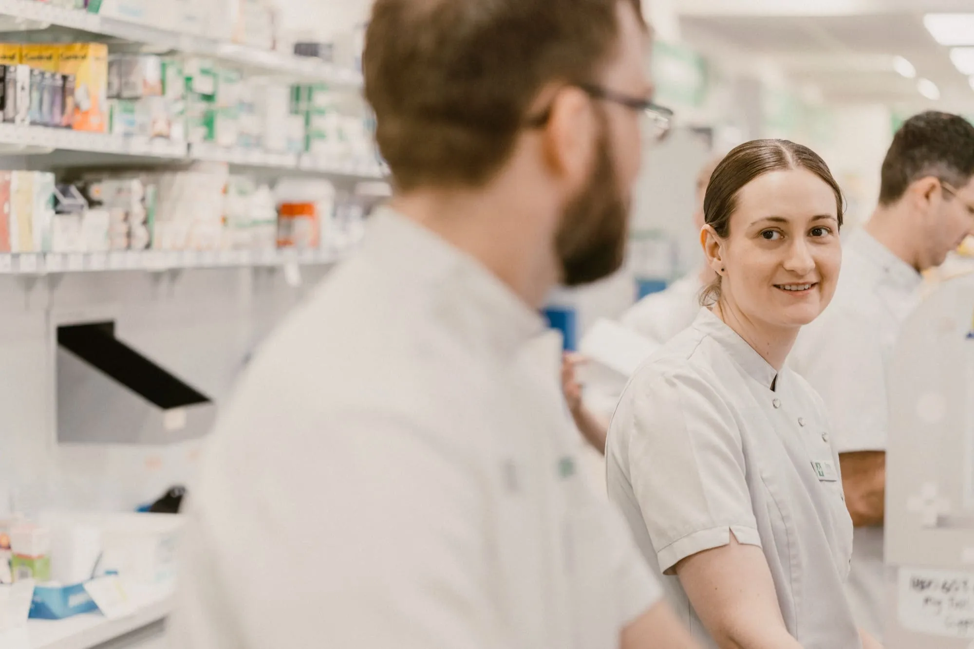 Pharmacists in white coats working behind the pharmacy counter with shelves of medication in the background.