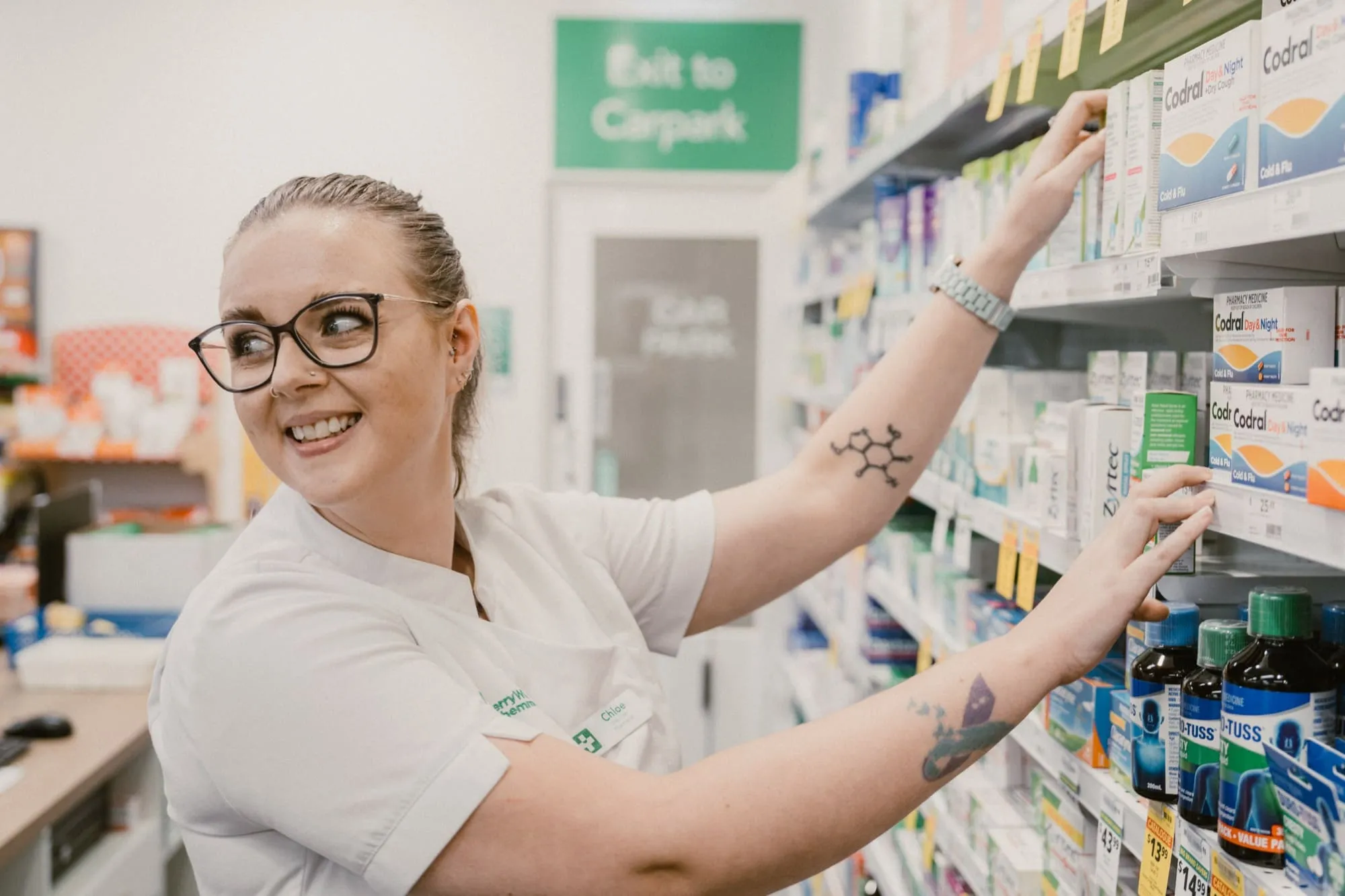 Smiling pharmacist with glasses and tattoos on arms arranging medicine boxes on a shelf in a pharmacy.