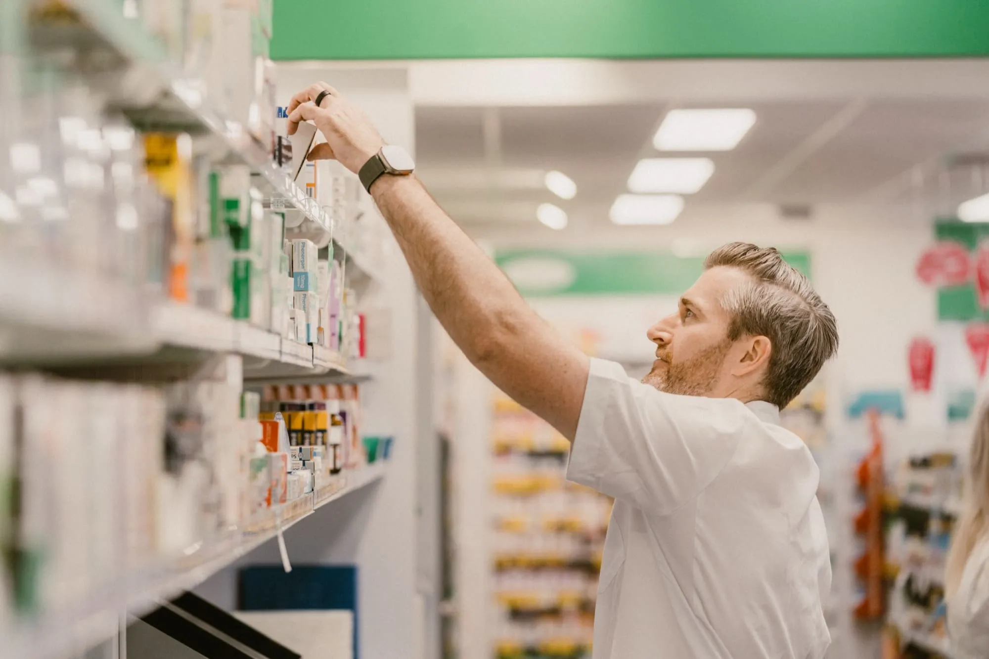 Male pharmacist in a white coat reaching for a product on a pharmacy shelf.