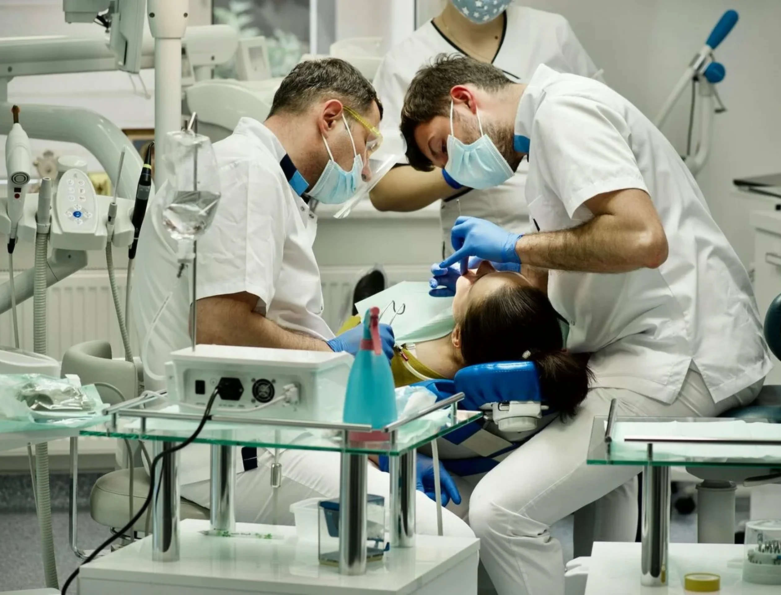 Two dentists wearing masks and gloves treating a patient reclining in a dental chair in a clinic.