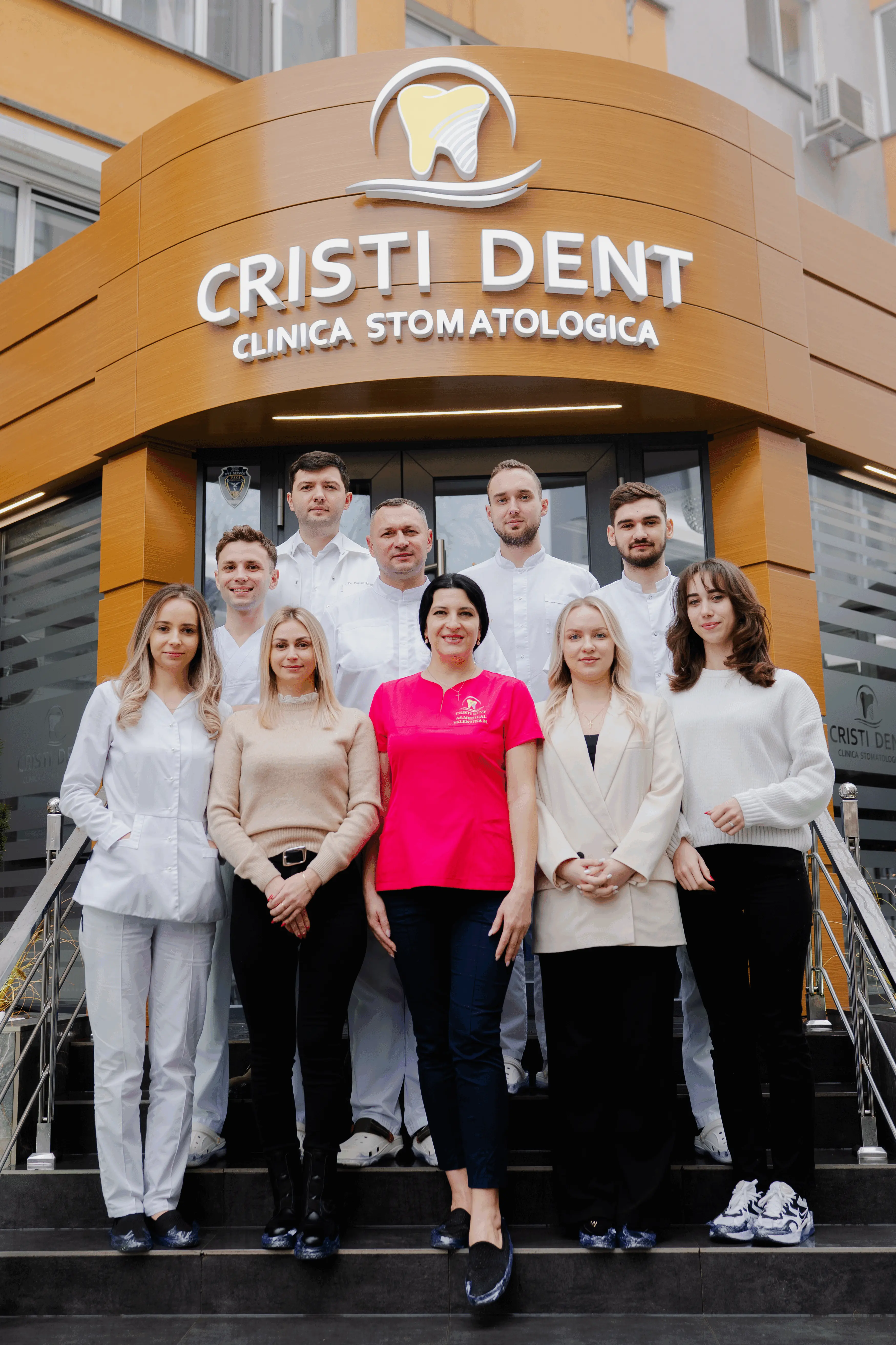 The CRISTI DENT dental clinic team stands on the steps in front of the clinic entrance. Nine staff members in medical attire pose beneath the “CRISTI DENT Clinica Stomatologica” sign, with a tooth-shaped logo displayed on the building facade.