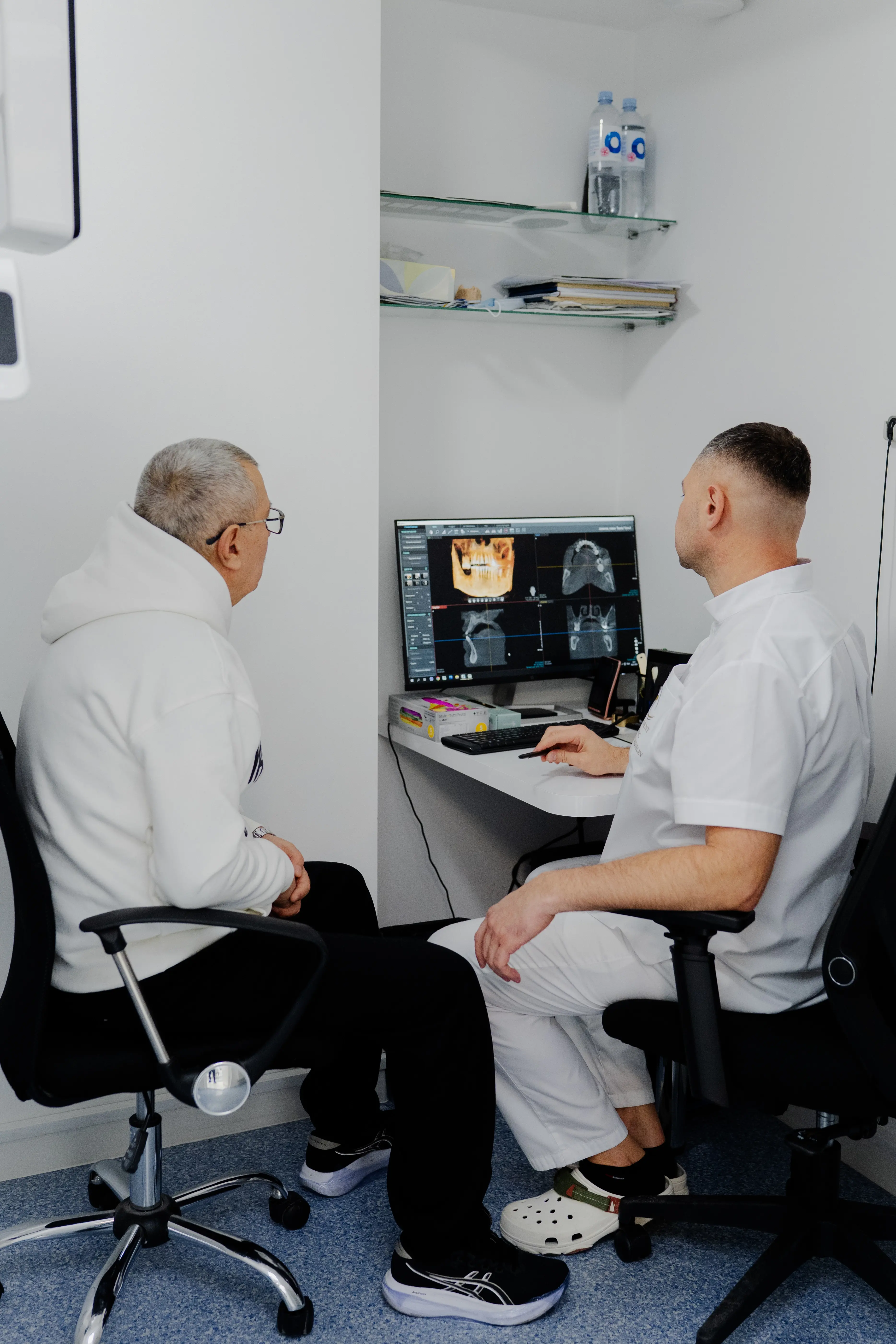 A dentist and an older male patient sit in a consultation room, reviewing 3D dental scan images on a computer monitor. The dentist points at the screen while explaining the treatment, and both are seated in office chairs in a clean, white clinical environment.