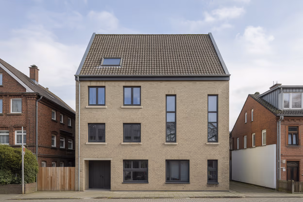 Modernes dreistöckiges Wohnhaus mit beiger Backsteinfassade und mehreren Fenstern unter bewölktem Himmel, flankiert von roten Backsteinhäusern.