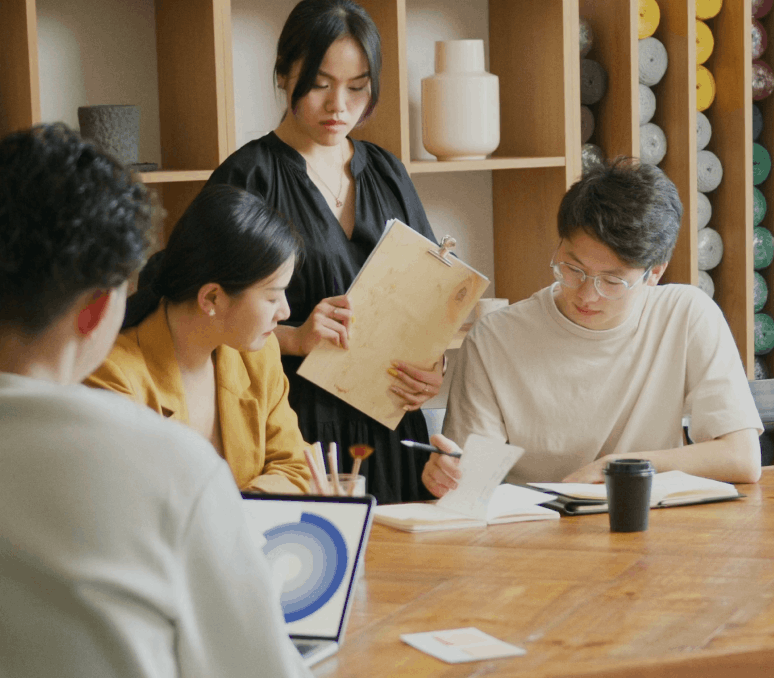 Four young adults collaborating around a wooden table with notebooks, a laptop showing a circular chart, and a coffee cup.