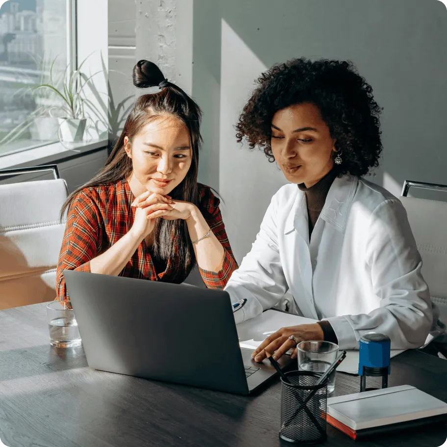 Two women, one in a plaid shirt and the other in a white coat, reviewing a laptop screen together at a desk with glasses of water and office supplies.