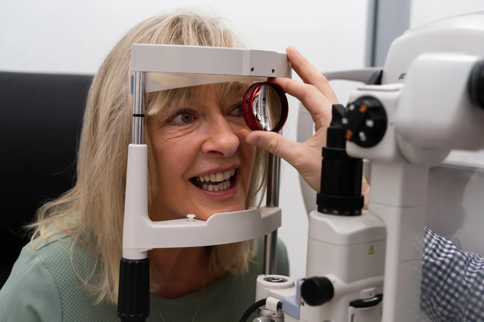 Older woman smiling during an eye exam with an ophthalmic device and a clinician holding a lens near her eye.