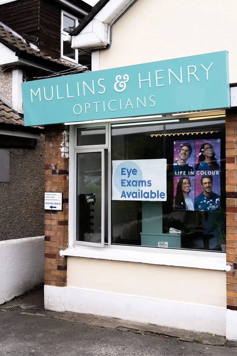 Storefront of Mullins & Henry Opticians with a blue sign and a window sign saying Eye Exams Available.