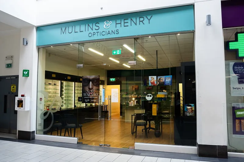 Front view of Mullins & Henry Opticians storefront with glass doors and displays of eyeglasses inside a shopping mall.