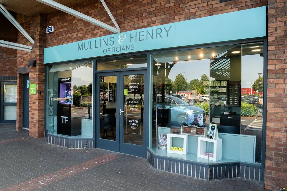 Exterior view of Mullins & Henry Opticians storefront with large windows displaying eyewear and a sign advertising hearing tests and ear wax removal service.