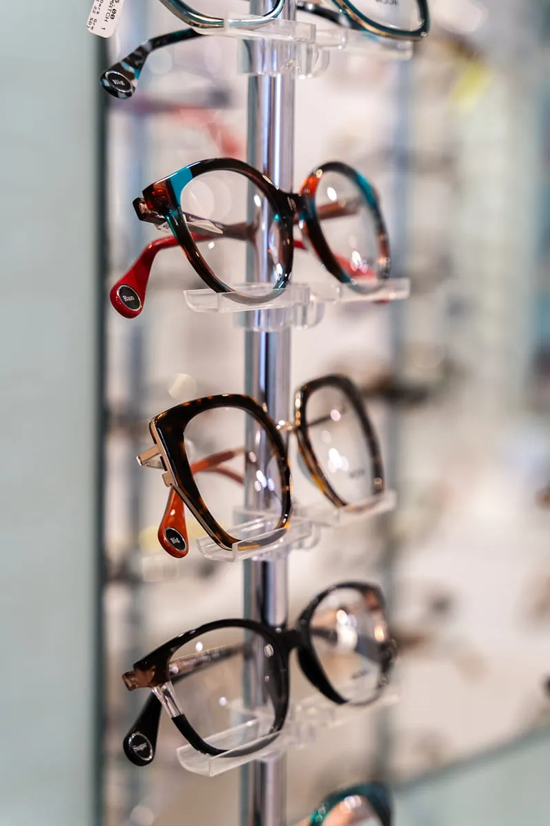 Four pairs of eyeglasses with different frame styles displayed vertically on a clear stand.