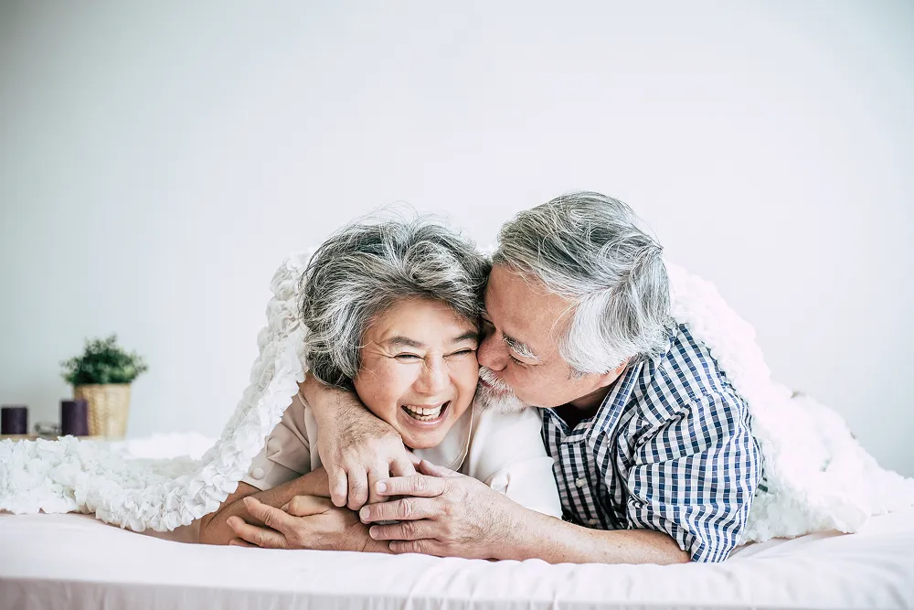 Elderly couple lying in bed smiling and embracing under a white blanket.