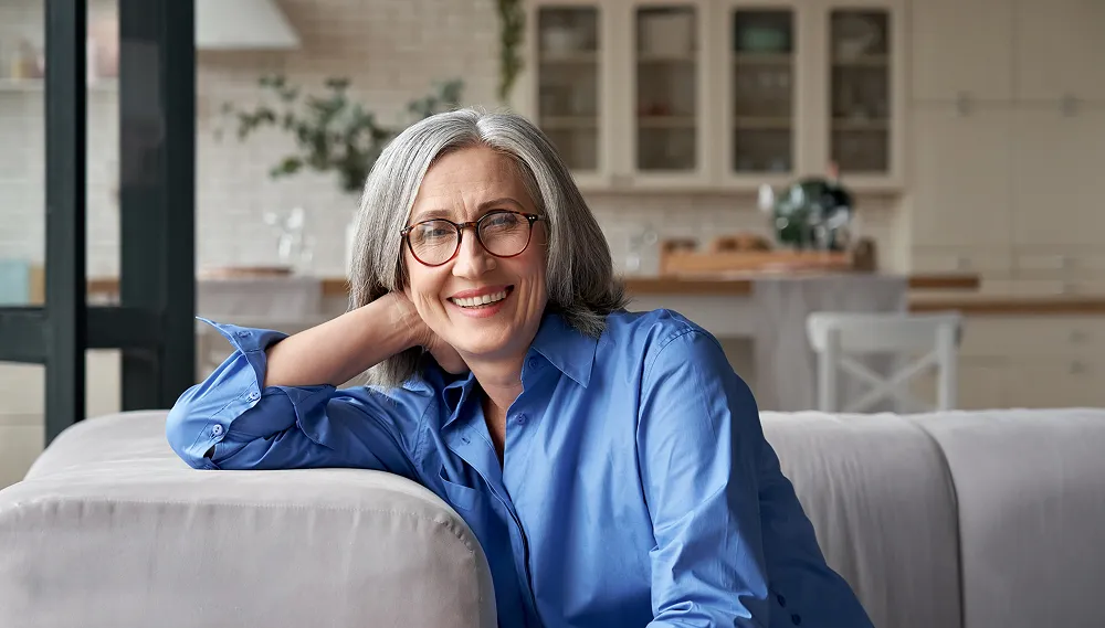 Smiling mature woman with gray hair and glasses leaning on a light gray sofa in a modern kitchen.