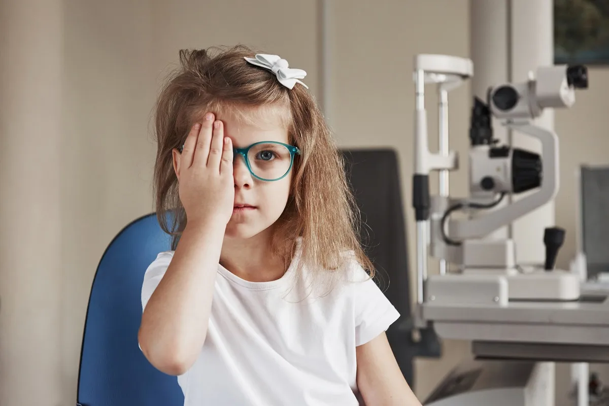 Young girl wearing glasses covering one eye with her hand during an eye exam in a clinic.