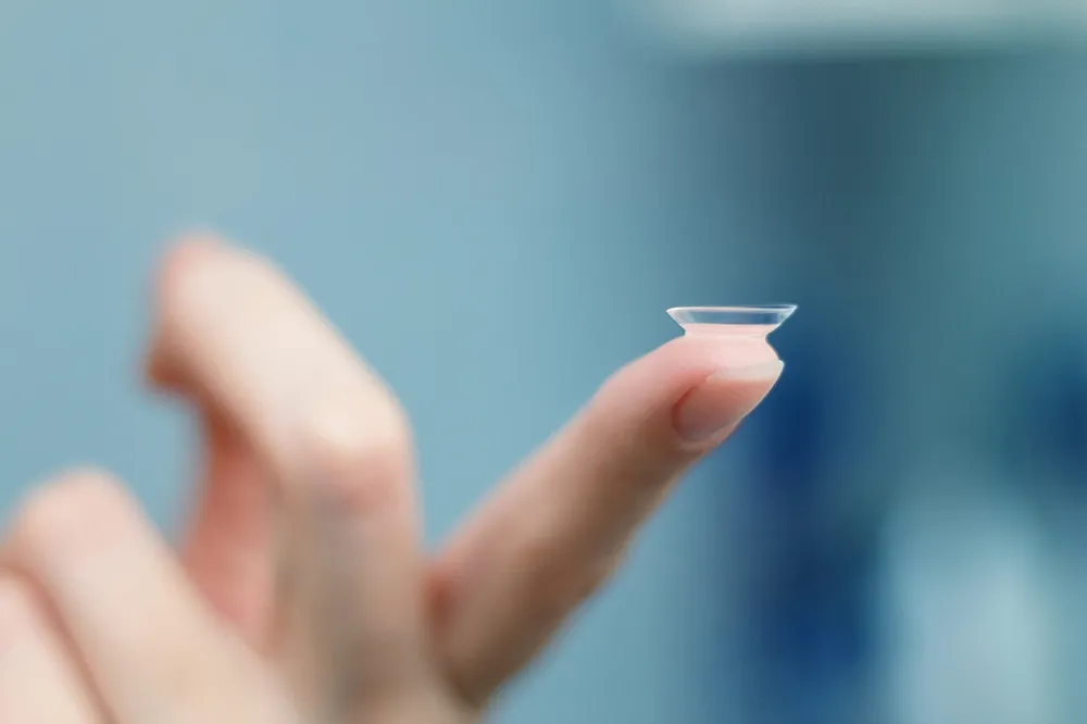 Close-up of a finger holding a transparent contact lens against a blurred blue background.