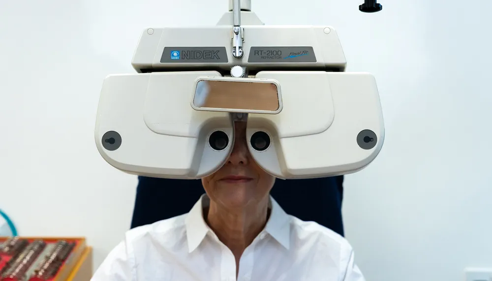 Person undergoing an eye exam using a Nidek RT-2100 refractor with face partially visible behind the machine.