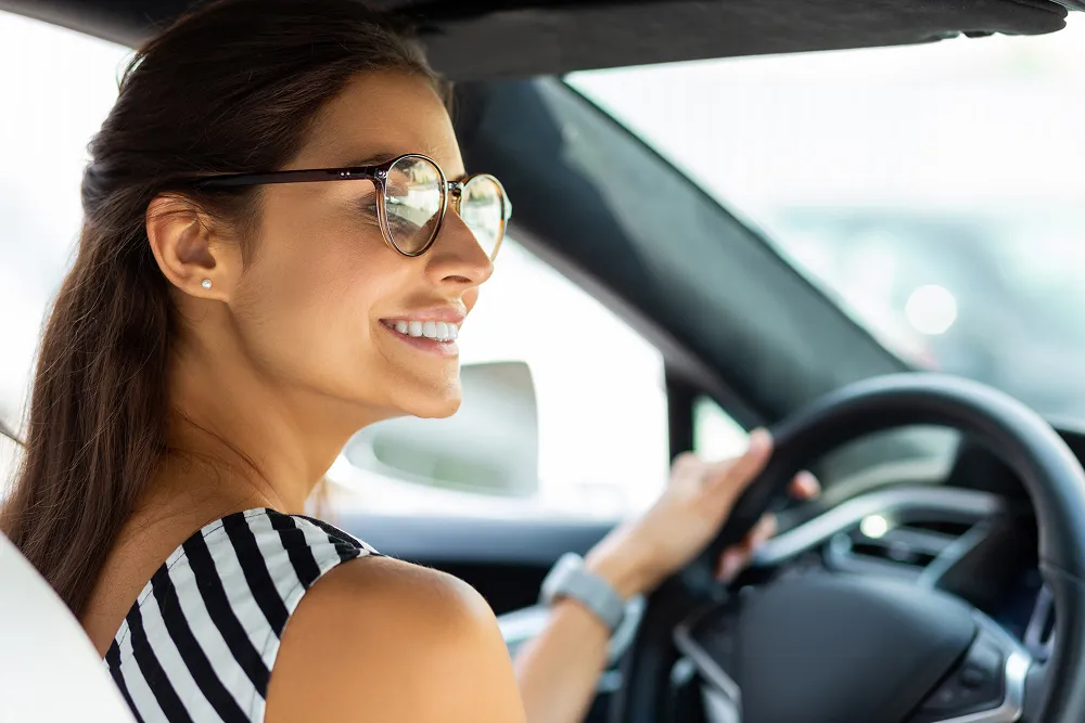 Smiling woman with long brown hair and glasses driving a car, looking over her shoulder.