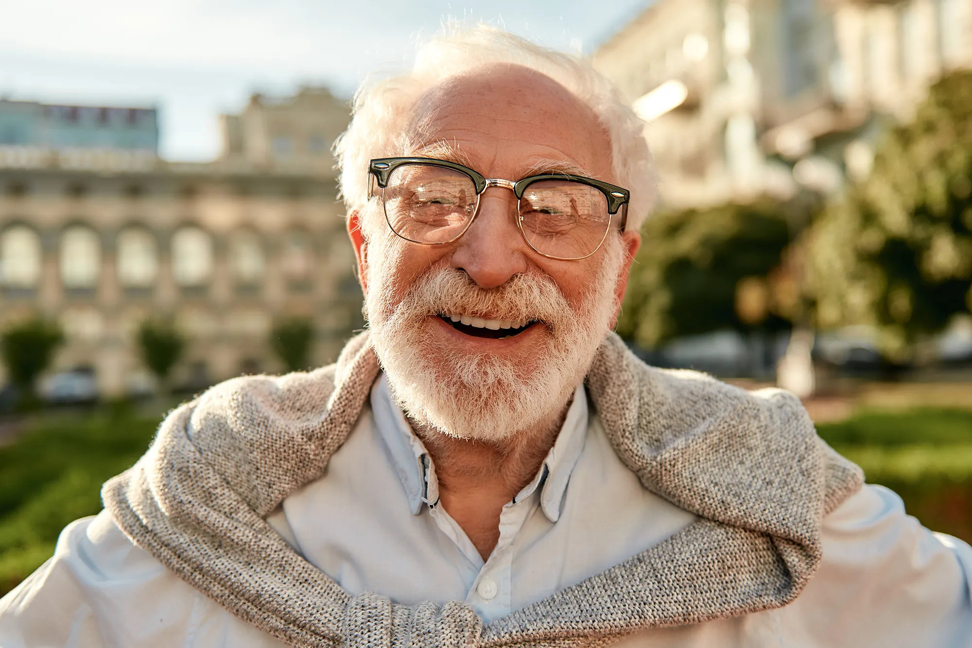 Smiling elderly man with white hair and beard wearing glasses and a light sweater draped over his shoulders outdoors.