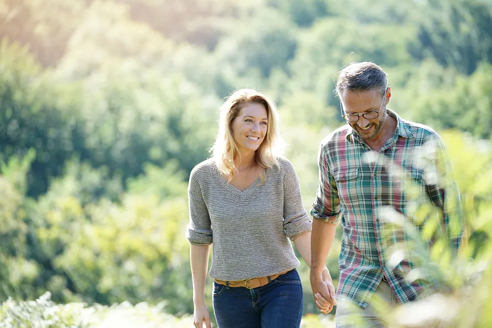 Smiling couple holding hands and walking outdoors with green trees in the background.