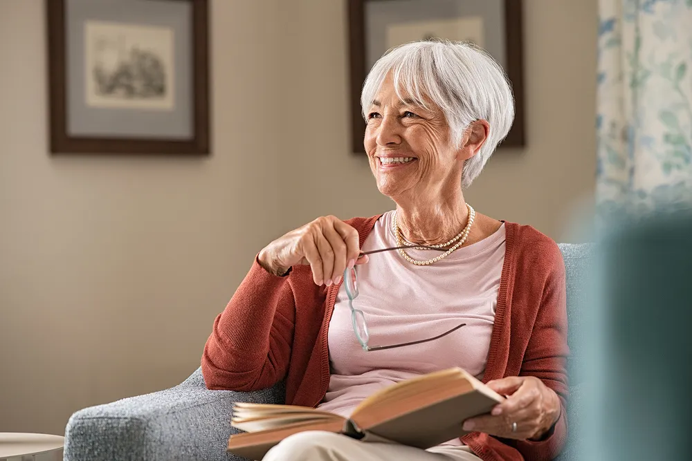 Smiling elderly woman with white hair holding eyeglasses and an open book while sitting on a gray armchair.