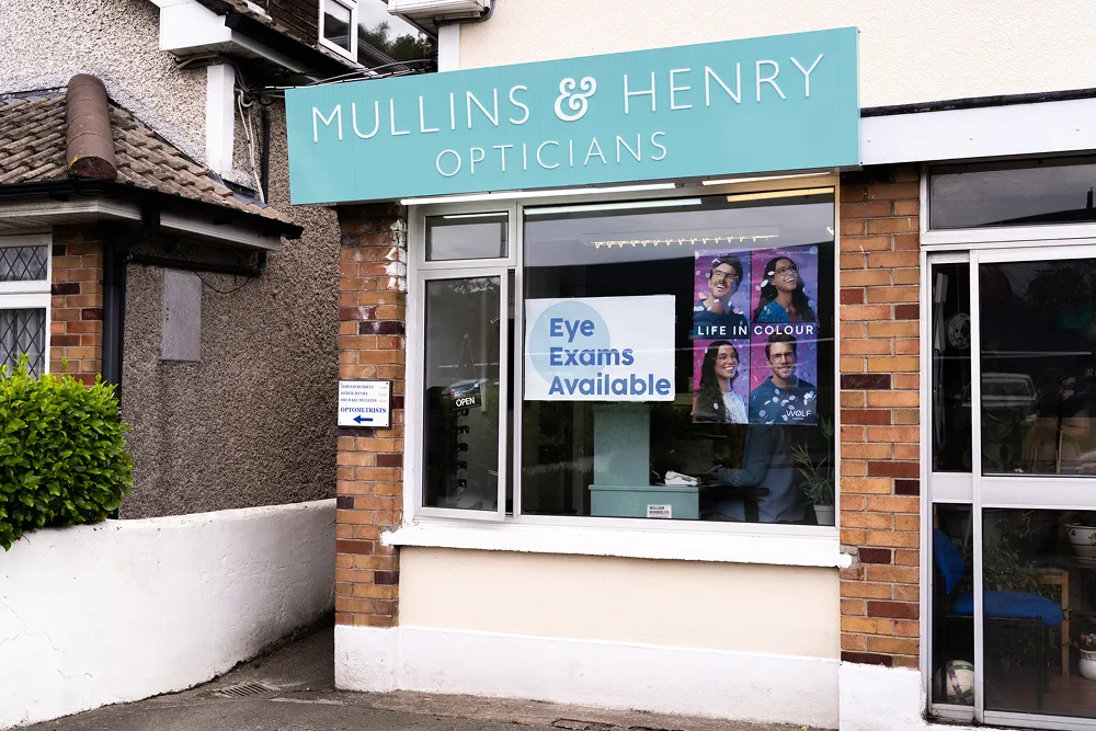 Shopfront of Mullins & Henry Opticians displaying signs advertising eye exams available and promotion posters with diverse models.