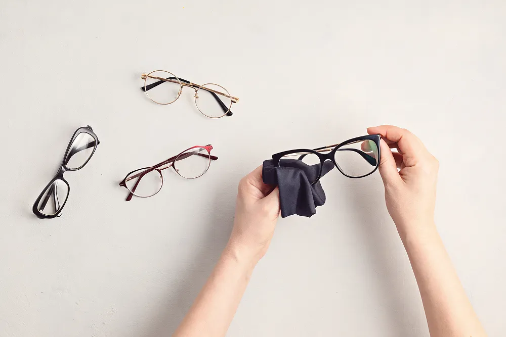 Hands cleaning black eyeglasses with a cloth, beside three pairs of eyeglasses on a light surface.
