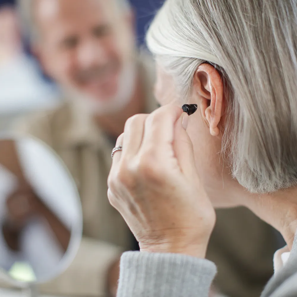 Elderly person with gray hair inserting a black hearing aid into their ear while looking into a mirror.