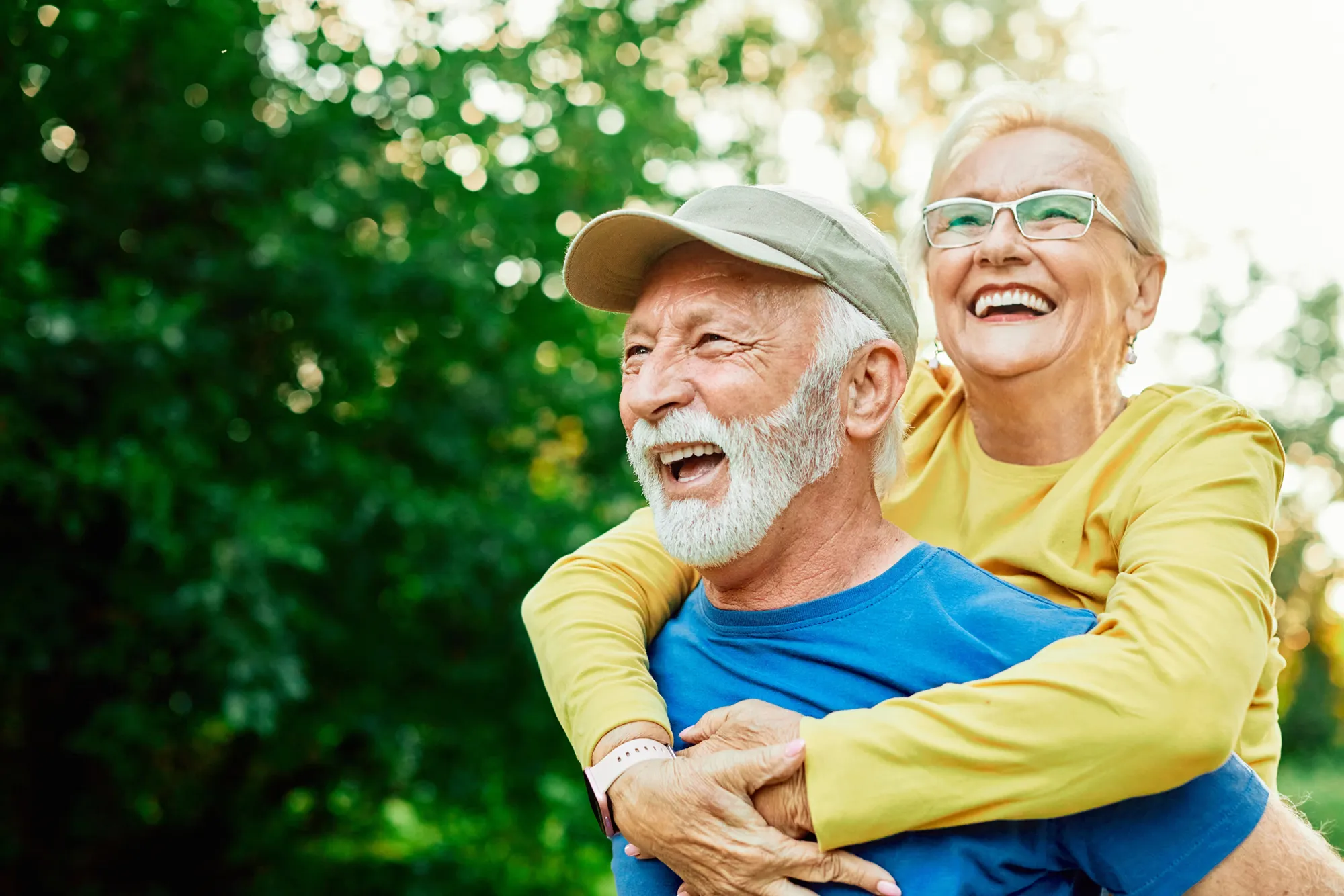 Smiling elderly couple outdoors, woman in yellow shirt hugging man in blue shirt and cap from behind.