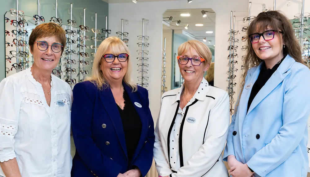 Four women wearing colorful eyeglasses and name tags, standing in an eyewear store with glasses displays behind them.