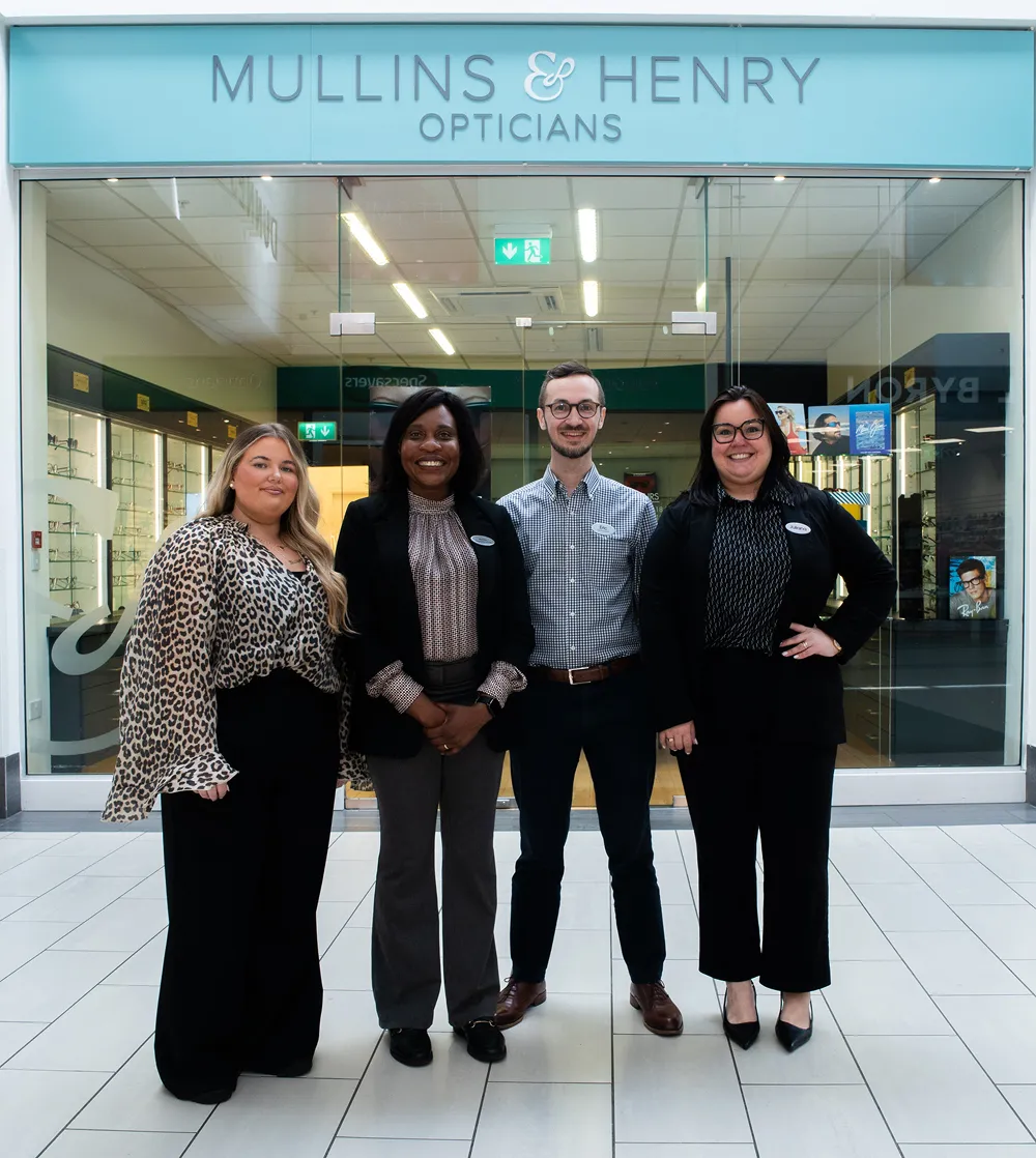 Four opticians standing in front of Mullins & Henry Opticians store, smiling and dressed in business casual attire.