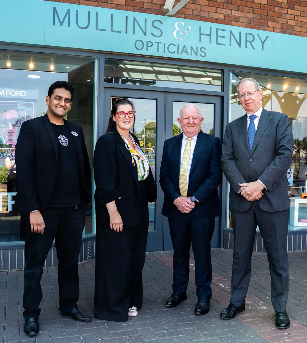Four professionally dressed people standing in front of Mullins & Henry Opticians store.