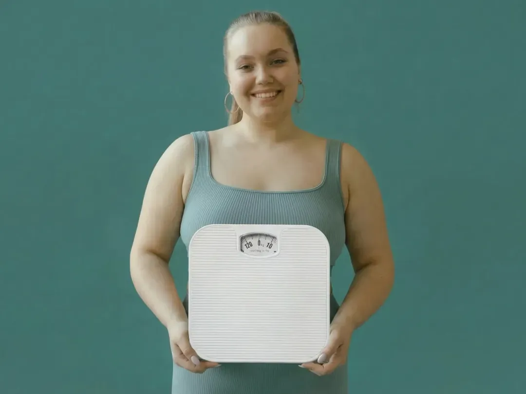 Une jeune femme souriante tient un pèse-personne blanc devant un fond bleu-vert, illustrant le bien-être et le contrôle du poids.