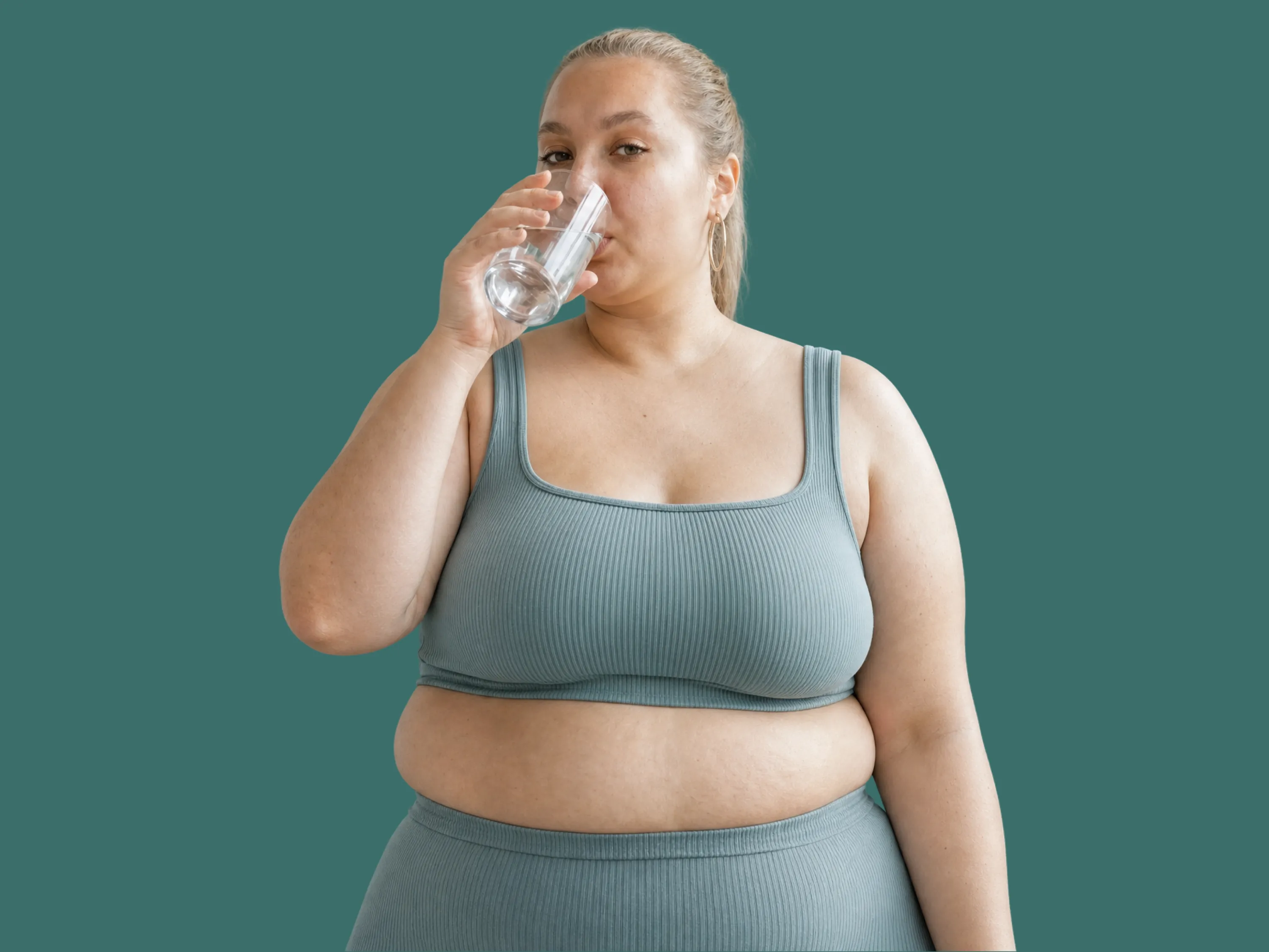 Une femme de forte corpulence aux cheveux blonds attachés boit un verre d'eau, vêtue d'un ensemble de sport bleu-vert, sur fond vert.