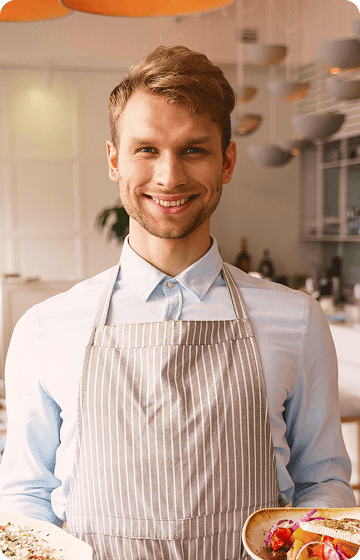 Усміхнений офіціант у світло-сорочці та смугастому фартусі тримає тарілки з їжею в ресторані.