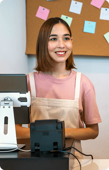 Усміхнена дівчина-касир в фартусі стоїть за касою з електронним терміналом.