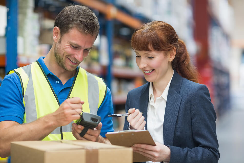 Warehouse worker wearing a high-visibility vest using a handheld scanner while a woman in a blazer holds a clipboard and smiles.