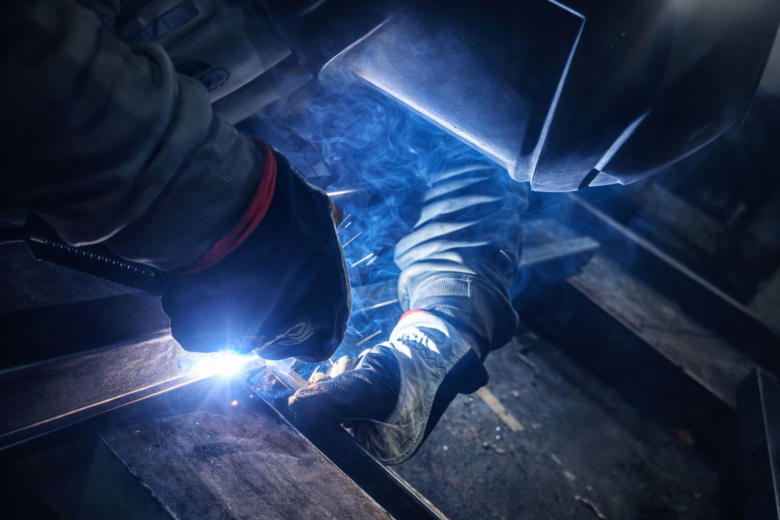 Worker welding metal with bright sparks and protective gloves and helmet in a dark workshop.