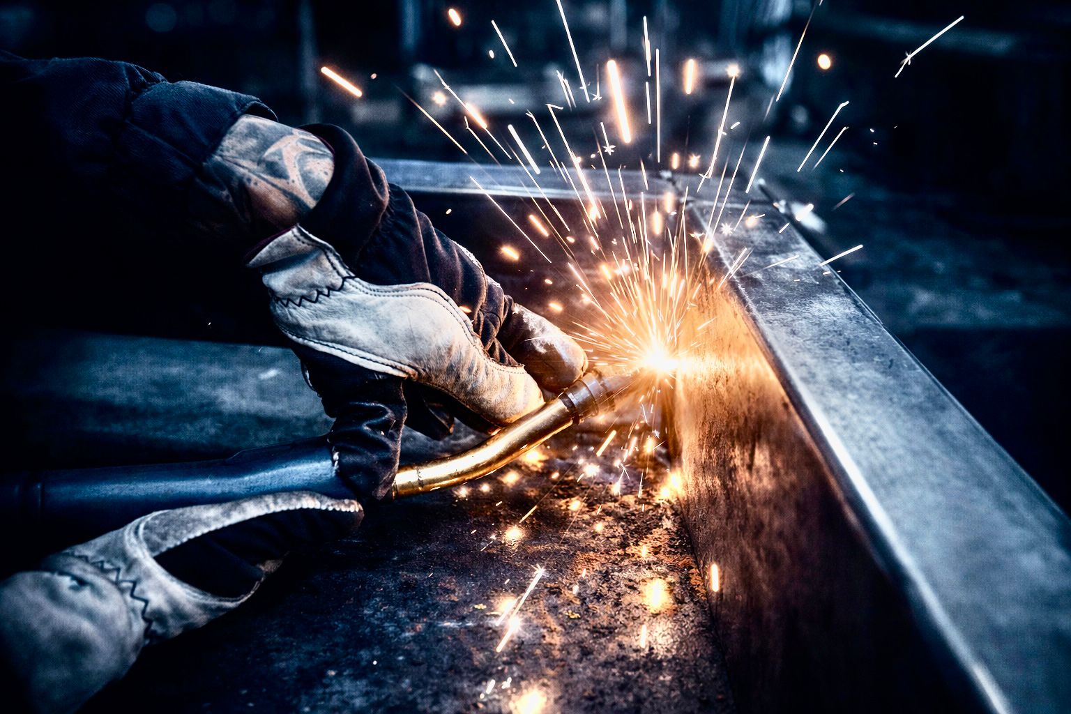 Gloved hand using a welding torch on metal, with sparks flying from the weld point.