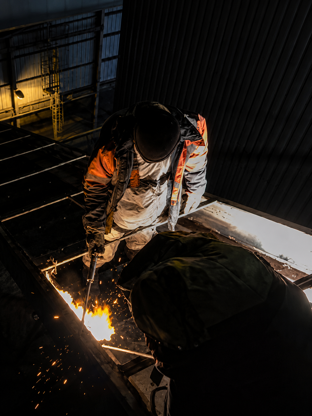 Two workers wearing protective gear using a torch to cut metal indoors with sparks flying.