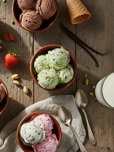 Bowls of chocolate, pistachio, and strawberry ice cream on a wooden surface with ice cream cone, vanilla pods, pistachios, a strawberry, two spoons, and a glass of milk.