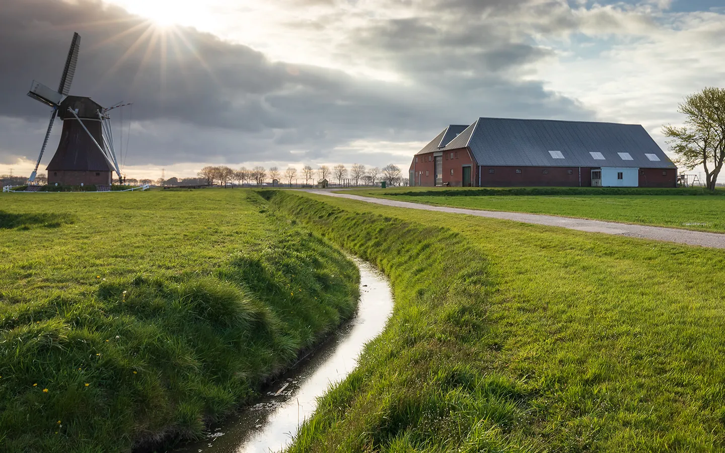 Rural scene with a traditional windmill, a barn, green grassy fields, and a narrow water canal under a partly cloudy sky with sun rays.