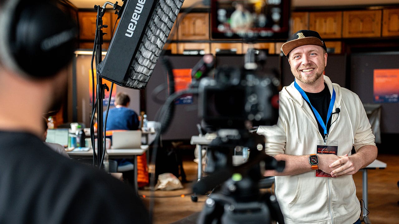 BENJAMIN HARRISON Global Head of R&D and Software Engineering from ELTEMATE mit Kappe und Schlüsselband wird für ein Eventvideo München in einem Innenbereich gefilmt und interviewt. Lichtanlage und Kamera sind zu sehen. Im Hintergrund arbeiten Menschen an Tischen mit Laptops und Postern.