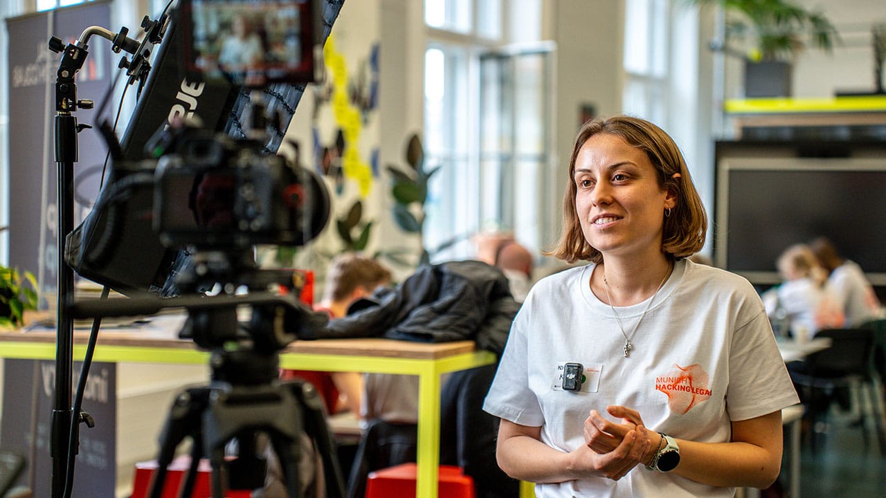 Eine Frau in einem hellgrauen T-Shirt und mit Mikrofon spricht in einer hellen, modernen Inneneinrichtung vor der Kamera, während im Hintergrund Menschen arbeiten und die Videoproduktion München für einen Unternehmensfilm vorstellt.