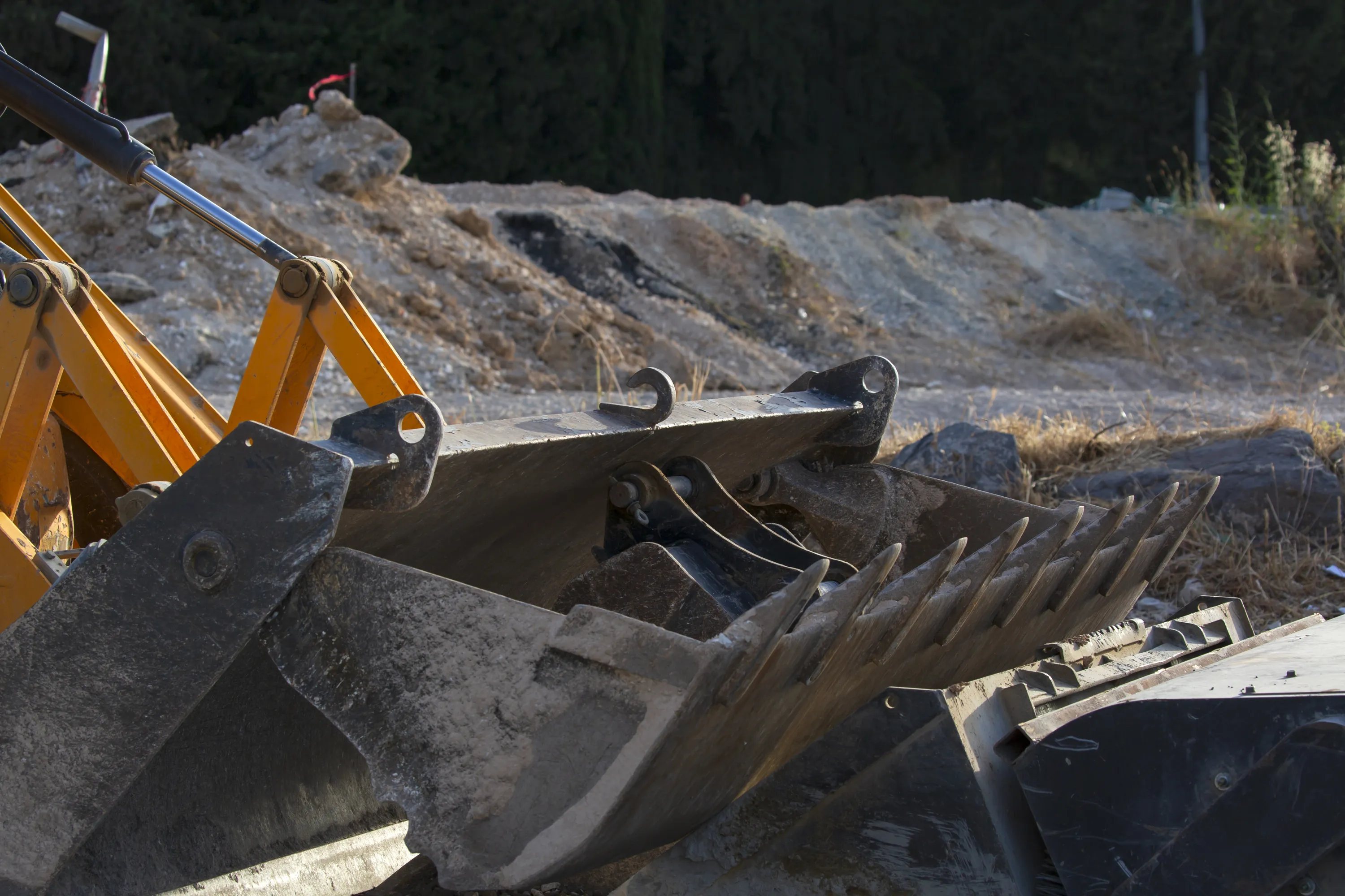 Lähikuva kaivinkoneen kauhoista, joissa on terävät hampaat ja kaksi koukkua, taustalla maaperää ja vihreää kasvillisuutta, valaistus on kirkas.