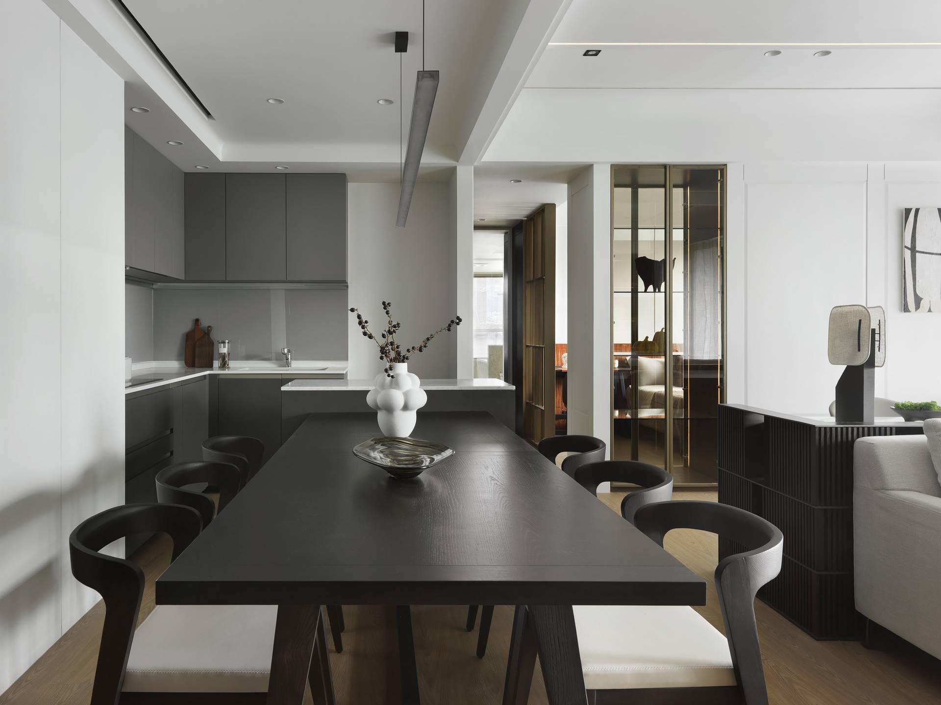 珥本設計｜台灣隱賦樣品屋 B5 餐廳場域：深色橡木長餐桌搭配黑色曲背餐椅一字排開，白色陶土花器插枯枝立於桌心，左側灰色極簡廚具與線形吊燈連續延伸，右側香檳金屬拉門與玻璃櫃隔間框入次臥景深