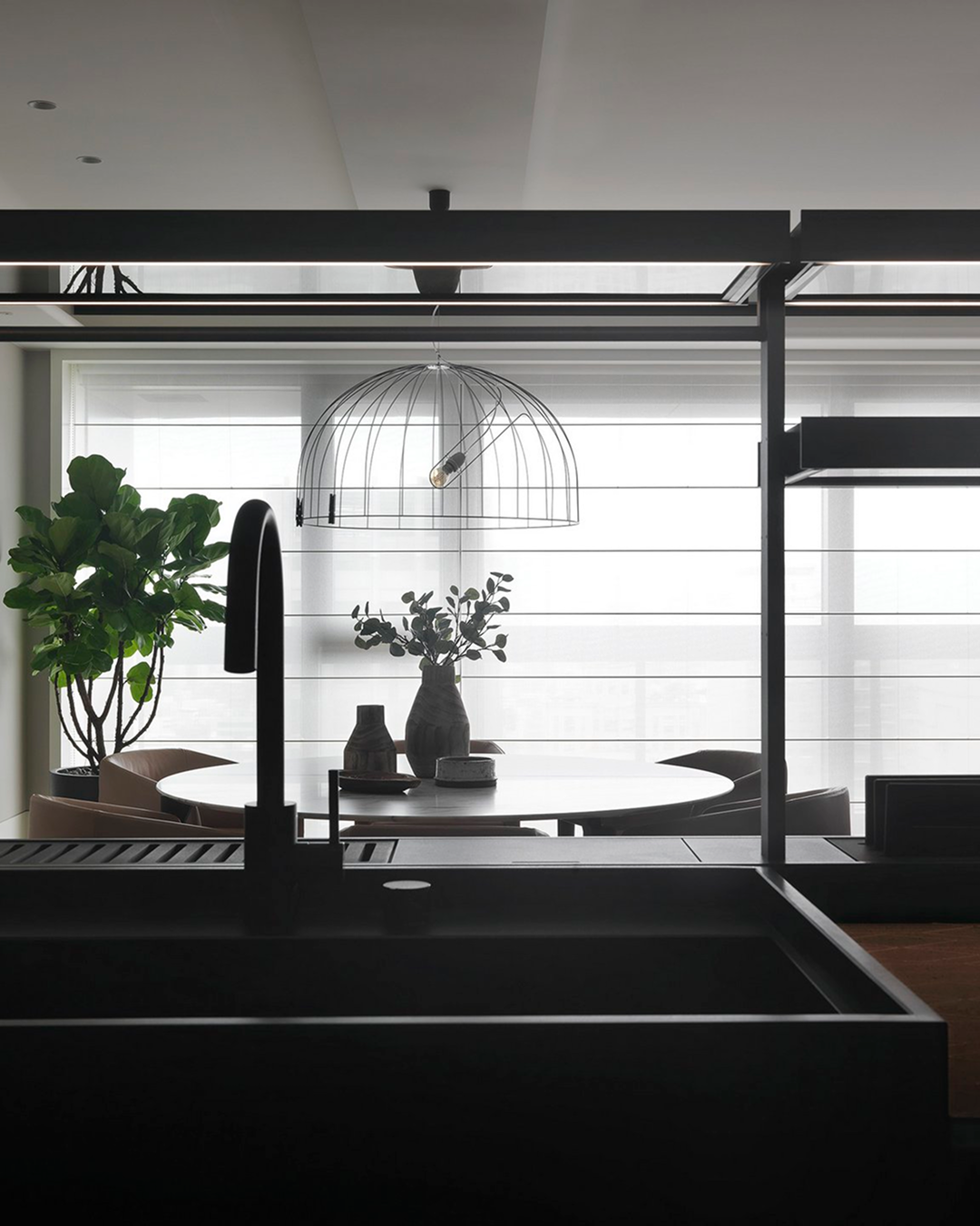珥本設計|丽格 BOFFI HOME 實品屋餐廚軸線:黑色方形水槽與弧形龍頭前景,圓形餐桌上灰陶花器與植栽,鳥籠造型金屬吊燈懸於上方,一側琴葉榕與百葉落地窗。