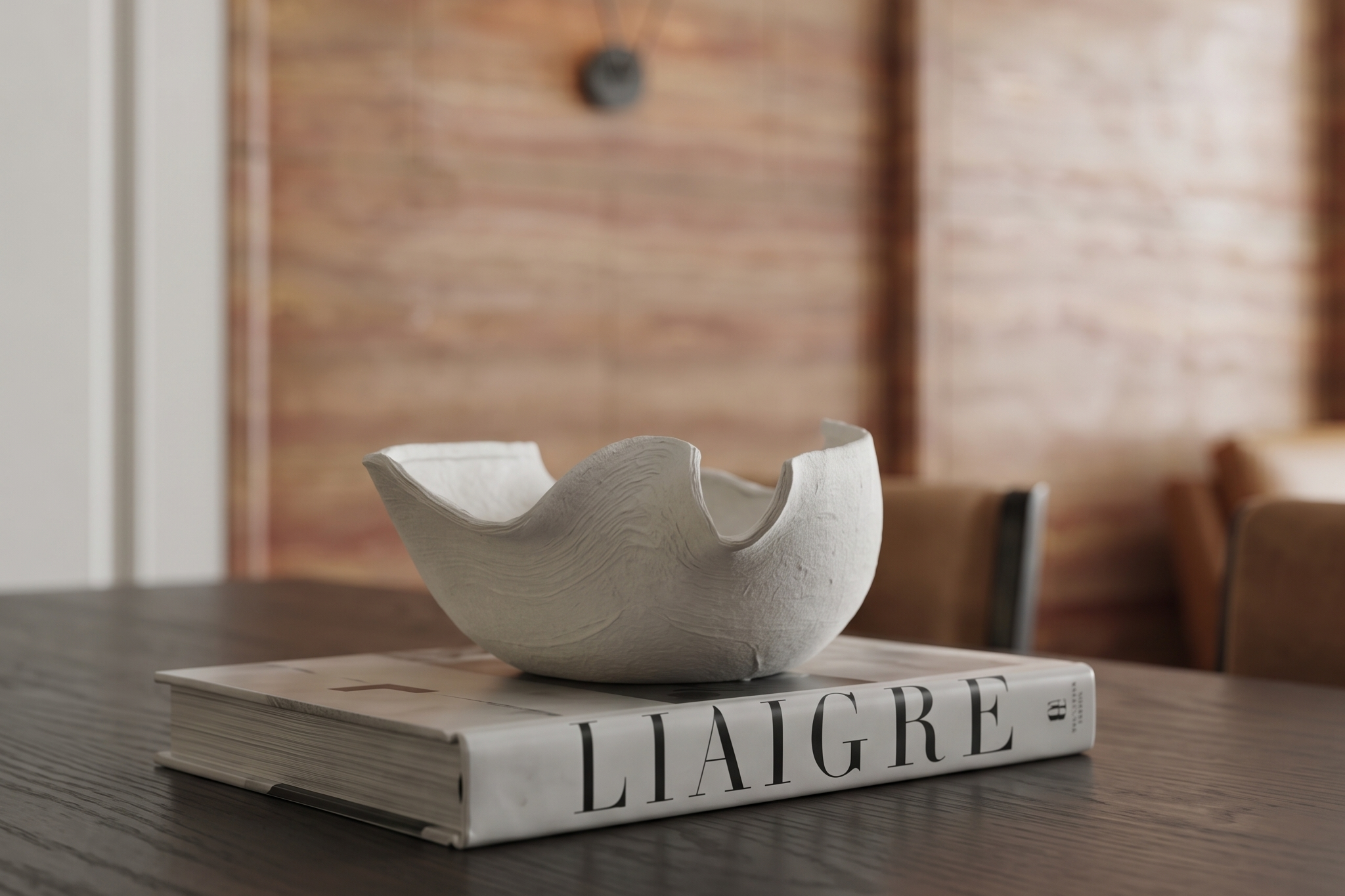 珥本設計｜藏賦樣品屋桌面静物：LIAIGRE 設計專書上置白色手作雕塑，下方深色橡木餐桌與背景虛化的紅色洞石紋理、駝色餐椅構成柔焦質地