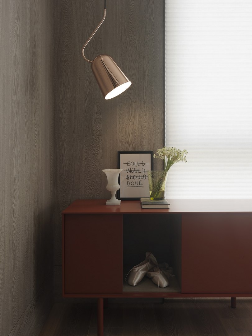 珥本設計|帝璟謙和樣品屋玄關端景:磚紅色 Muuto Reflect 邊櫃承載白色石雕甕、黑框標語畫與玻璃花瓶,深灰褐色木皮直紋牆為底,玫瑰金銅色垂吊燈懸於側,右側白色橫紋風琴簾漫射日光