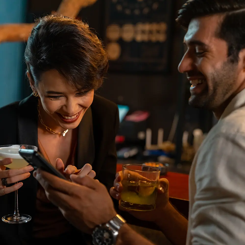 A man and woman smiling and looking at a smartphone while holding drinks at a bar.