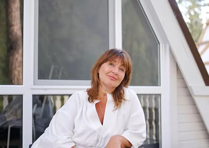 Woman with brown hair wearing a white blouse sitting in front of a large window on a porch.