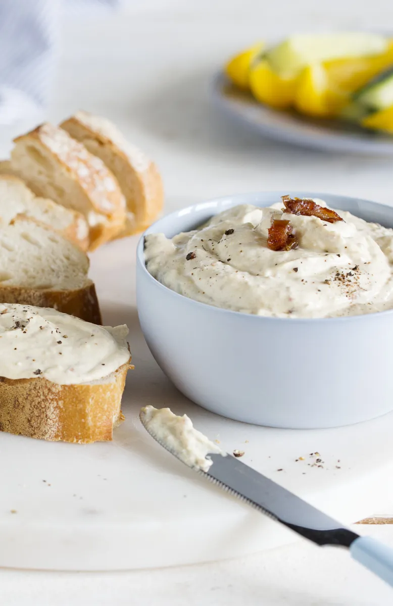 Bowl of creamy hummus garnished with two cherry tomato halves and sprinkled with chili flakes, next to a spoonful of hummus and a small bowl of crushed chili flakes on a white wooden surface.